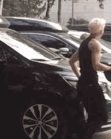 a man in a black tank top is standing next to a black car in a parking lot