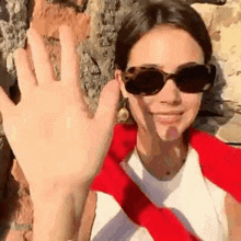 a woman wearing sunglasses and a red sweater is waving her hand .