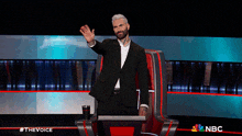 a man in a suit waves while sitting in front of a podium that says the voice on it