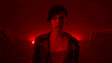 a woman in a studded leather jacket stands in a dark room with red lights behind her