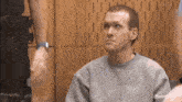 a man in a gray sweater is sitting in a room with a police officer in the background .