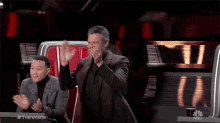 a man in a suit is applauding while a man in a black turtleneck sits behind him on the voice