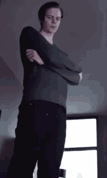 a man in a grey sweater and black pants is standing with his arms crossed in front of a window .