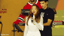 a woman in a white dress is singing into a microphone while a man in a red hockey jersey stands behind her .