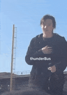 a man in a black jacket is standing in front of a blue sky holding his hands to his chest .