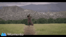 a woman in a dress is running through a grassy field with mountains in the background and the word momento in the corner