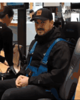 a man wearing a black jacket and a hat is sitting in a car seat with a blue harness