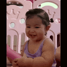a baby girl in a purple tank top is smiling in front of a pink fence