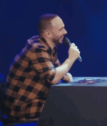 a man in a plaid shirt is singing into a microphone while sitting at a table