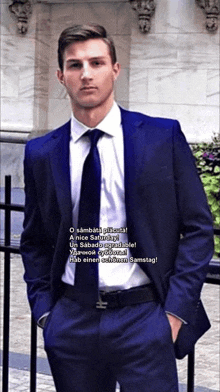 a man in a blue suit and tie is standing in front of a fence with his hands in his pockets