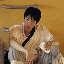 a man wearing a white shirt with a dog embroidered on it sits on the floor holding a cup