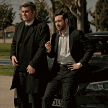 two men standing next to a black car with license plate number 4y75
