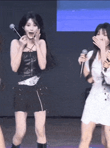 a woman in a black top is singing into a microphone while a woman in a white dress covers her mouth