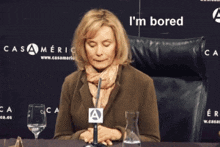 a woman sits at a table with a microphone and a sign that says i 'm bored