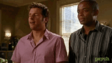 two men in striped shirts are standing next to each other in front of a window with blinds .