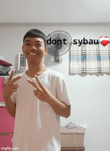 a young man stands in front of a wall with a fan and a sign that says dont sybau