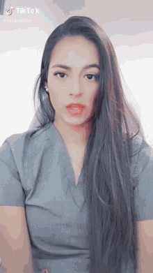 a woman with long dark hair is wearing a grey scrub top and has a tiktok logo above her