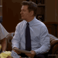 a man in a blue shirt and tie is sitting on a couch with peacock written on the bottom right