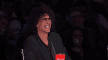 a man with curly hair is sitting in front of a microphone and laughing while holding a cup of soda .