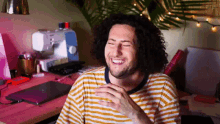 a man with curly hair is laughing in front of a sewing machine and a laptop .
