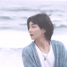 a young man wearing a blue cardigan and a white t-shirt is standing on the beach .