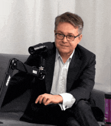 a man in a suit sits on a couch in front of a rode microphone