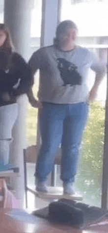 a man in a white shirt with a black cat on it is standing in front of a window in a room .