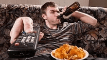 a man is sitting on a couch eating chips and drinking beer .