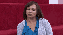 a woman in a blue shirt and gray sweater is sitting on a red carpet .