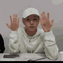a young man wearing a white hat and glasses is sitting at a table with his hands up .