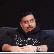 a man wearing a black sweatshirt with a horse on it sits on a couch