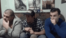 three men are sitting in a room with posters on the wall and playing a video game .