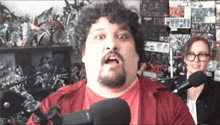 a man in a red shirt is talking into a microphone while a woman in glasses looks on .