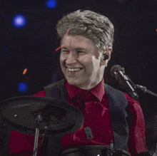 a man in a red shirt is playing drums and smiling in front of a microphone