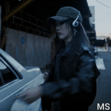 a woman wearing headphones and a hat is standing next to a car with the letters ms on the bottom right