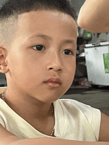 a young boy wearing a white tank top and a necklace looks at the camera