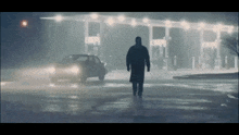 a man in a trench coat walks in front of a car at a gas station