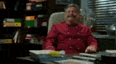 a man in a red chef 's jacket sits at a desk in front of a window