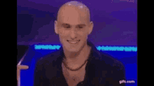 a bald man is smiling and clapping his hands while sitting in front of a blue background .