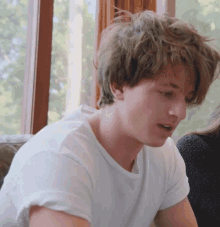a young man with messy hair wearing a white shirt is sitting in front of a window