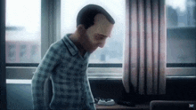 a man in a plaid shirt is standing in front of a window with a kc logo on the bottom right