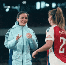 a woman in a blue jacket talks to another woman in a red and white jersey with the number 2 on it