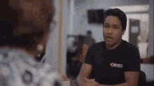 a man in a black shirt is sitting at a table talking to another man .