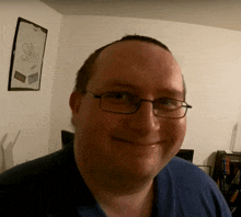 a man wearing glasses and a blue shirt smiles in front of a framed picture that says ' a sakko '