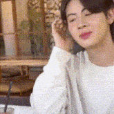 a young man in a white shirt is sitting at a table with his eyes closed and talking on a cell phone .