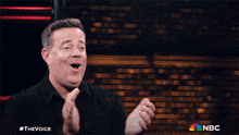 a man applauds in front of a brick wall with the nbc logo in the corner