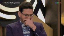 a man wearing glasses and a purple suit is covering his mouth in front of a masterchef logo
