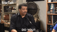 a man in a black shirt is sitting in front of a microphone and says do you