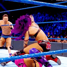 a woman with purple hair is in a wrestling ring with a man standing behind her .
