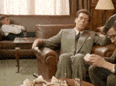 a man in a suit sits in a chair while another man lays on a couch